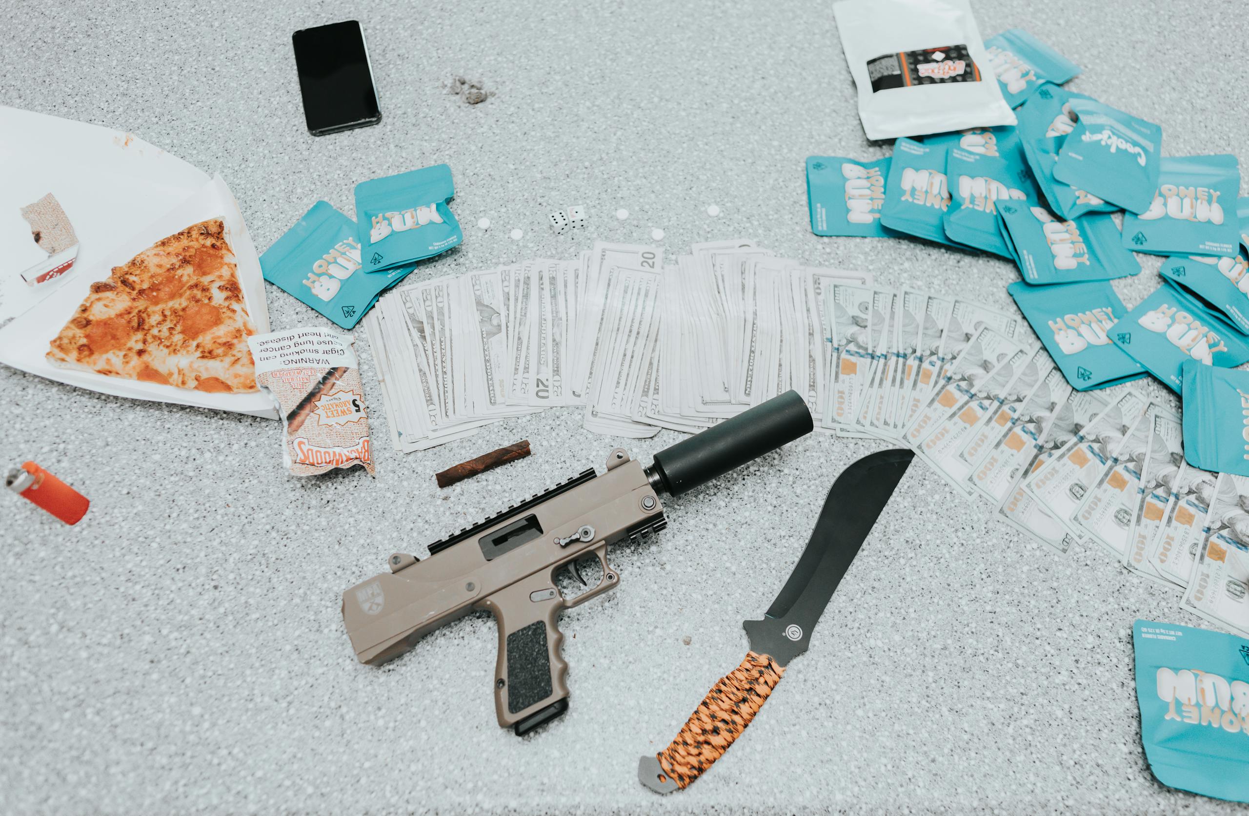 Close-up view of cash, a firearm, and a knife on a table with a slice of pizza.