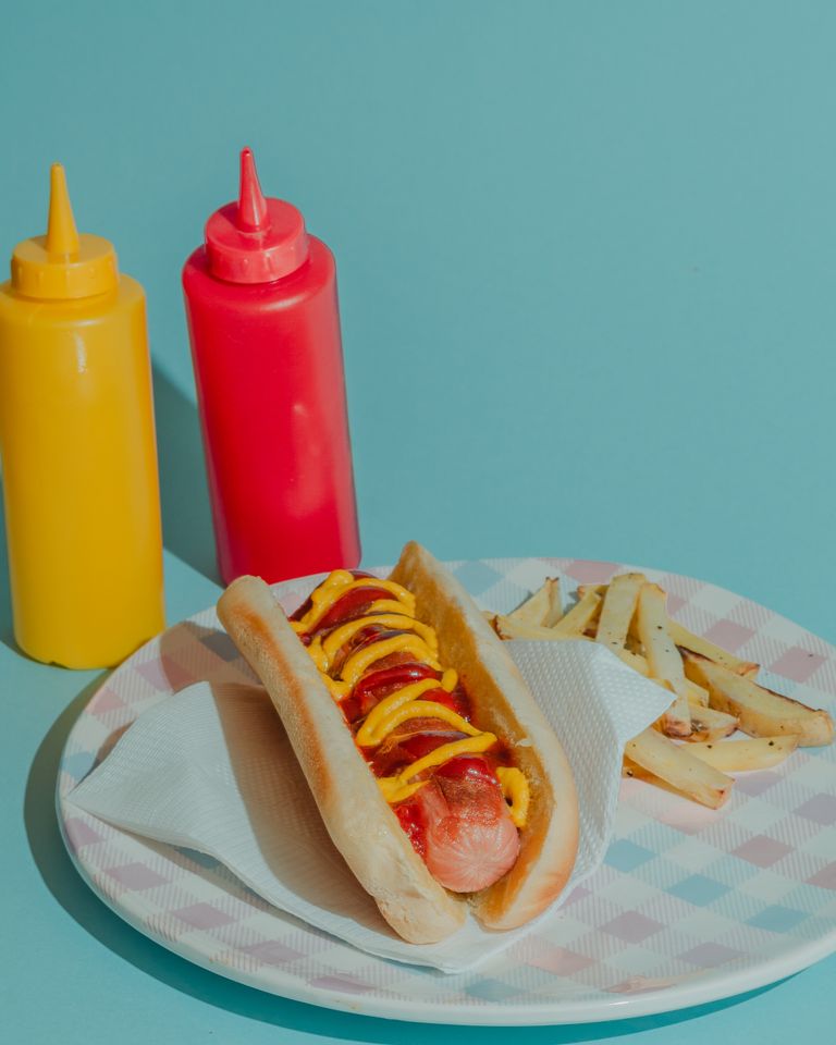 Colorful hot dog and fries setup with mustard and ketchup bottles on a pastel background.