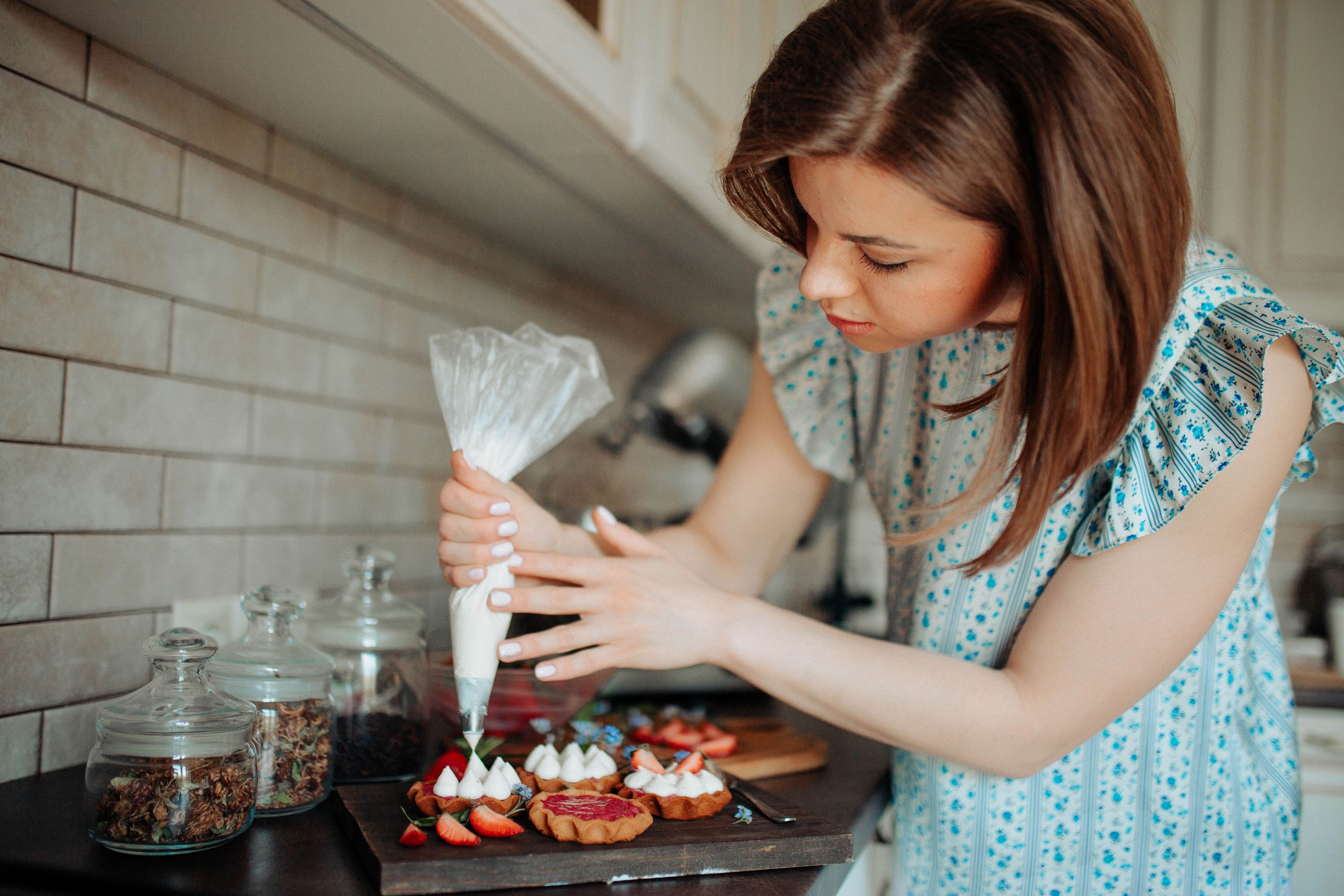 Concentrated young female in casual clothes with pastry bag decorating cookies with cream and strawberries on table in light kitchen