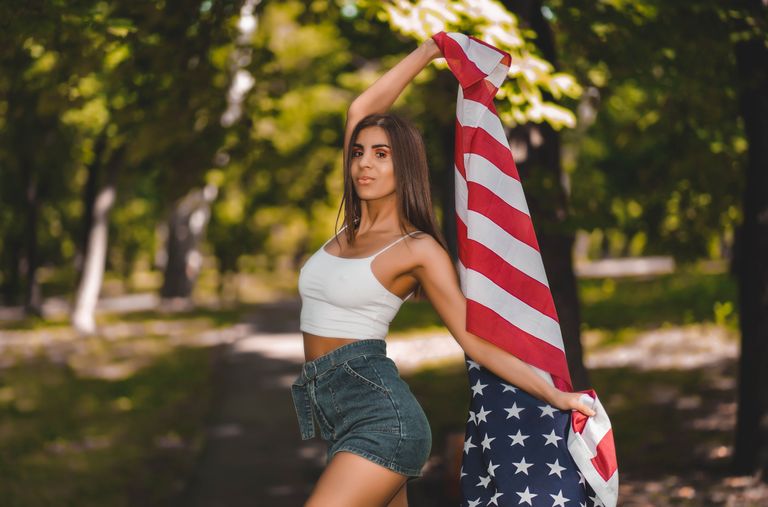 A confident woman with an American flag is posing outdoors in a park.