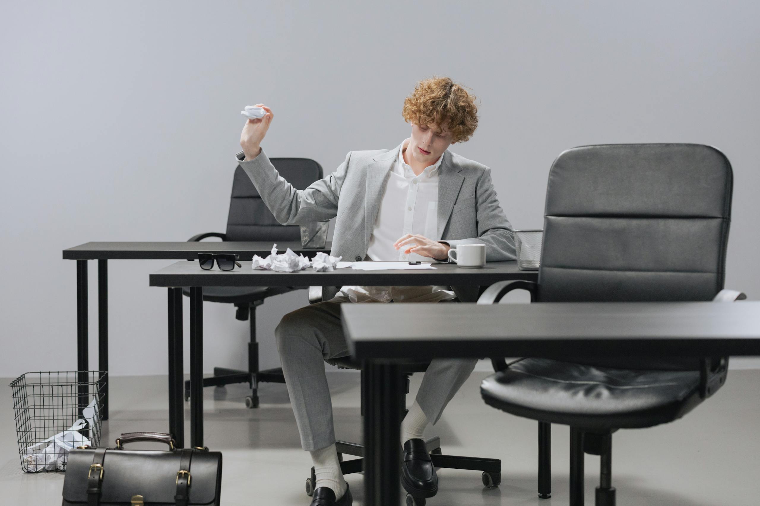 Confident young man in gray suit tossing crumpled paper in modern office setting.