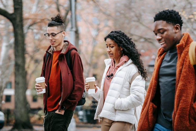Confident young multiracial friends in outerwear and backpacks talking while strolling in city street near trees with paper cups with drink and earphones in daytime in park