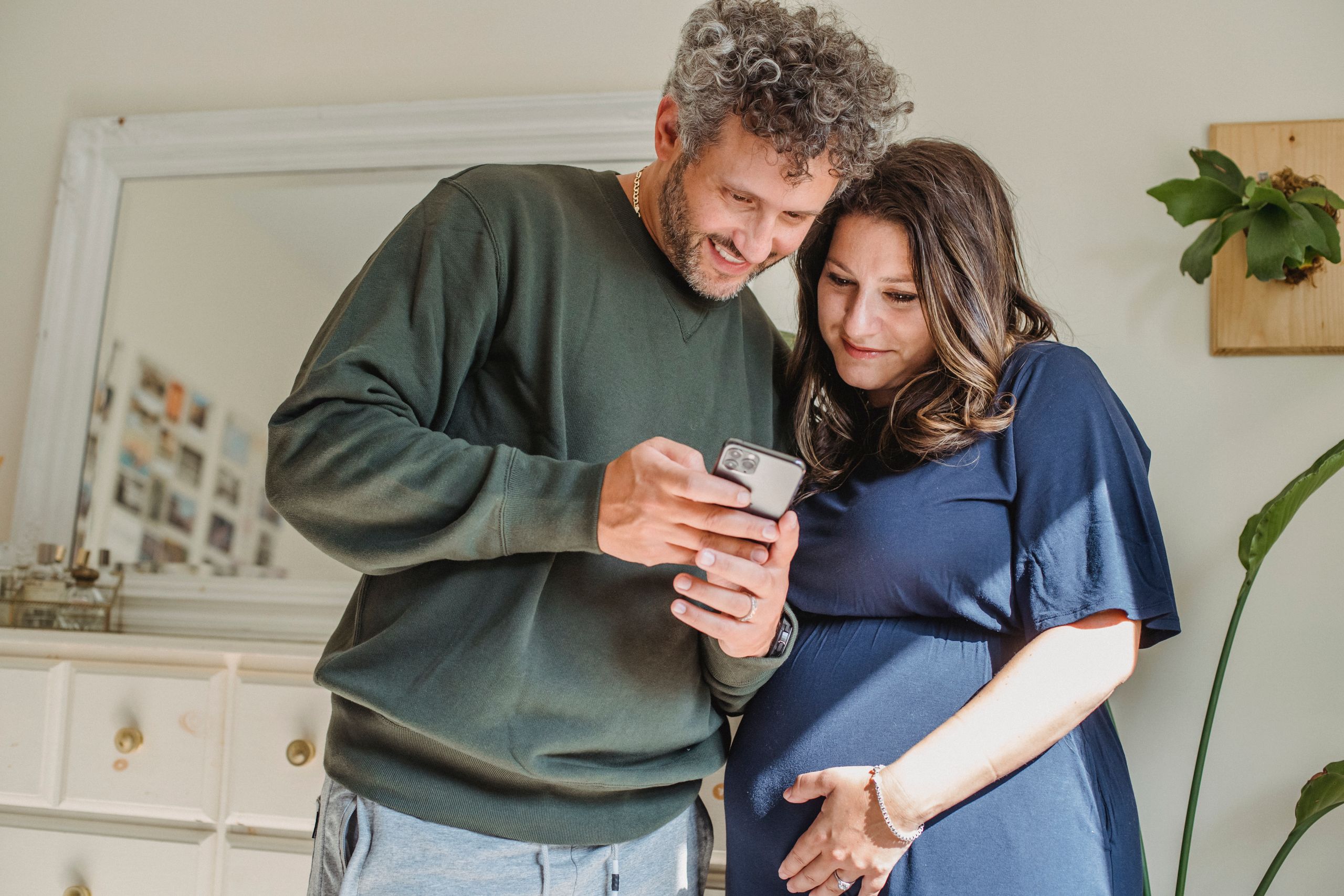 Content bearded male showing cellphone to feminine expectant woman caressing tummy in house room in sunlight