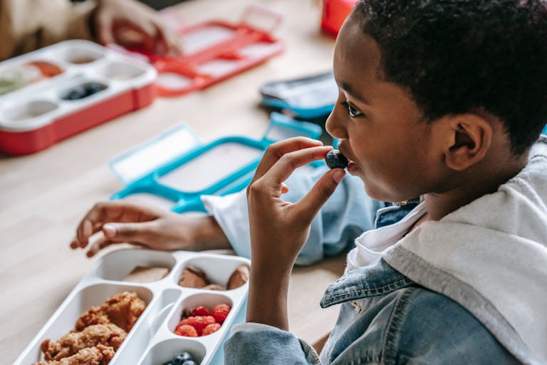 A cropped African American child in casual clothes sitting at table with plastic lunch box while having snack