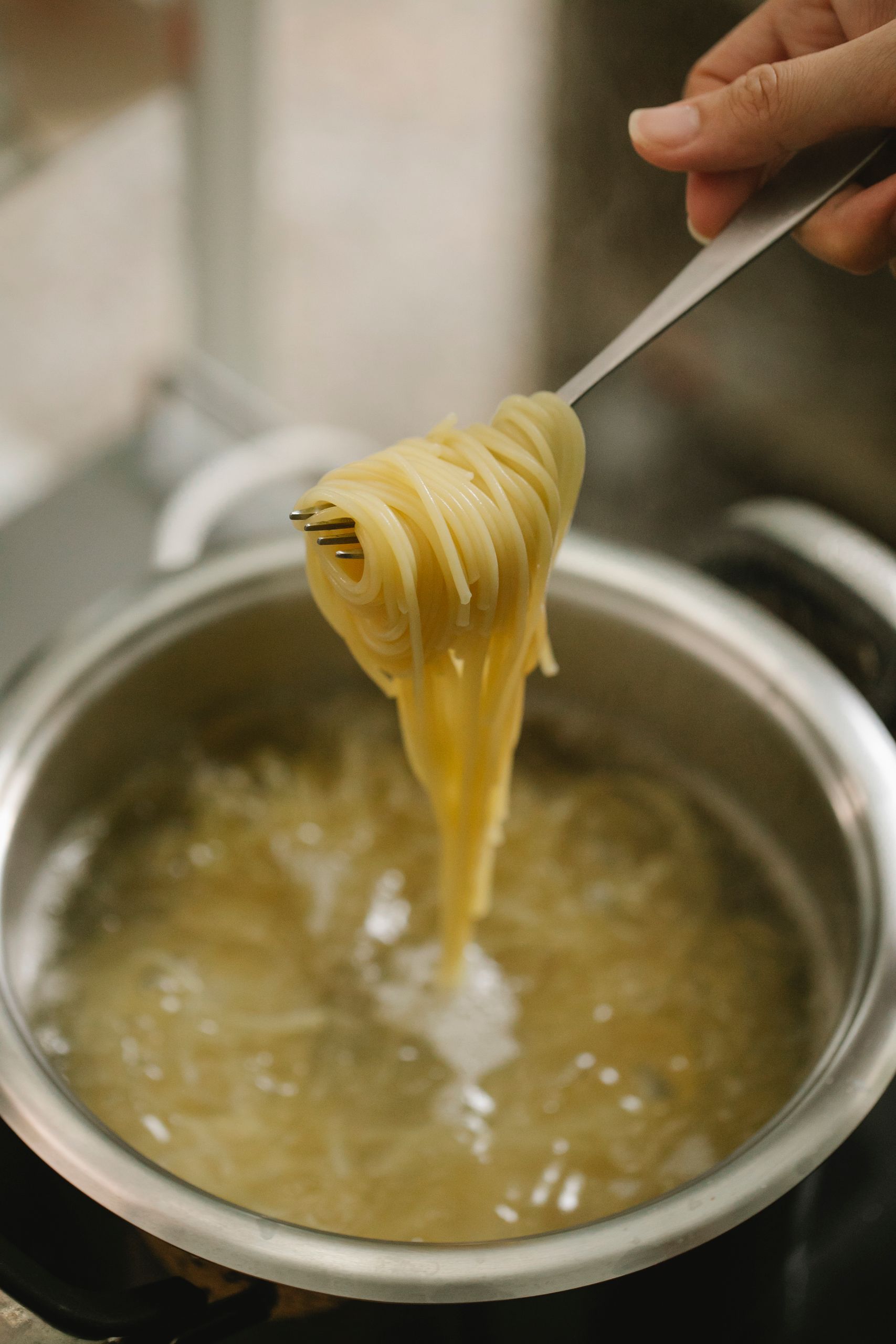 Crop anonymous chef showing fork with spaghetti above saucepan with boiling water while cooking pasta in kitchen