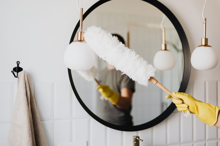 Crop anonymous female housewife in rubber gloves dusting modern lamp with soft brush against mirror while cleaning stylish bathroom