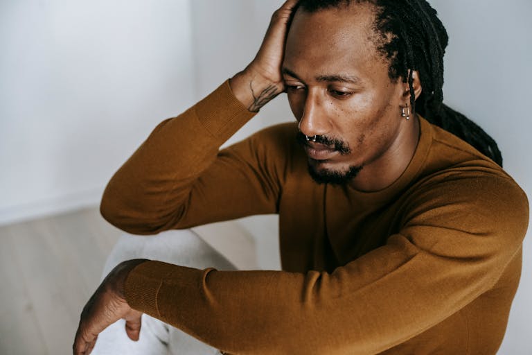 Crop young hopeless African American male with piercing touching head and looking down in house