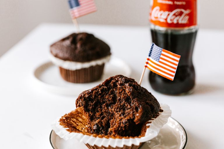 Delicious bitten chocolate cupcakes decorated with cocktail flag of USA and served on white table with bottle of coke