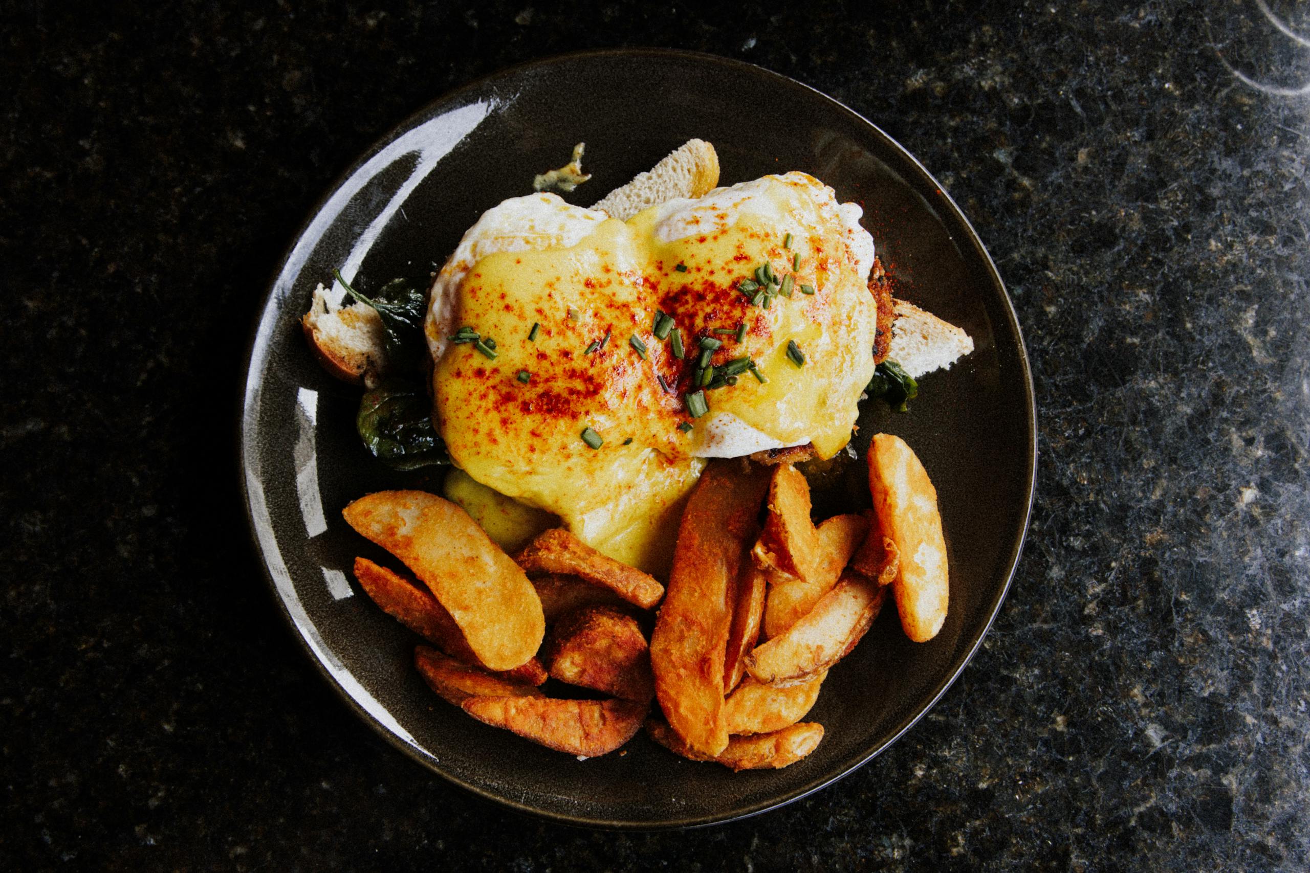 Delicious Eggs Benedict with creamy sauce and crispy potato wedges served on a dark plate.