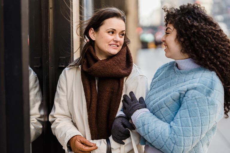 Delighted young diverse female friends in trendy warm clothes smiling and looking at each other while chilling on city street on cold day