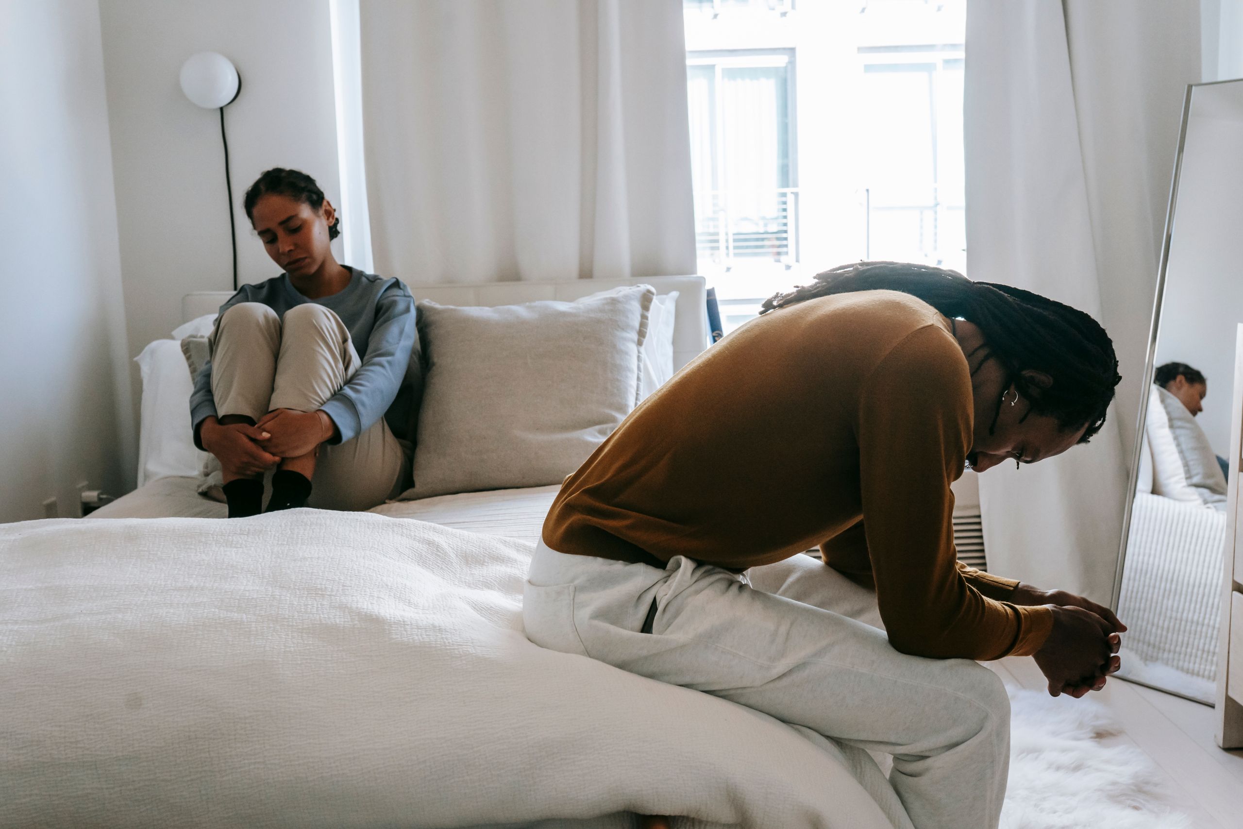 Discontented black man with clasped hands sitting on bed near upset girlfriend during quarrel in apartment