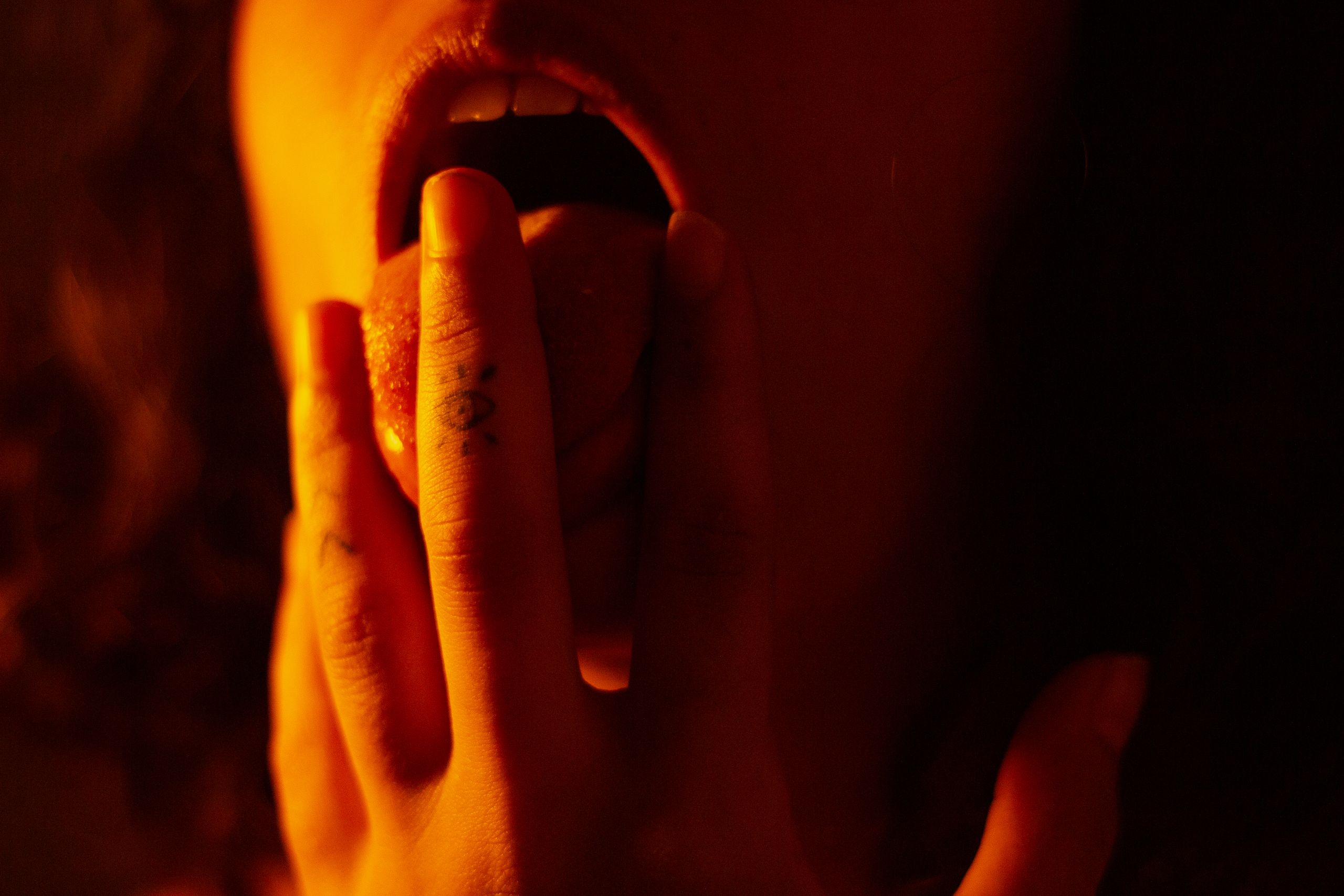 Dramatic close-up of fingers with a tattoo touching a tongue under warm lighting.