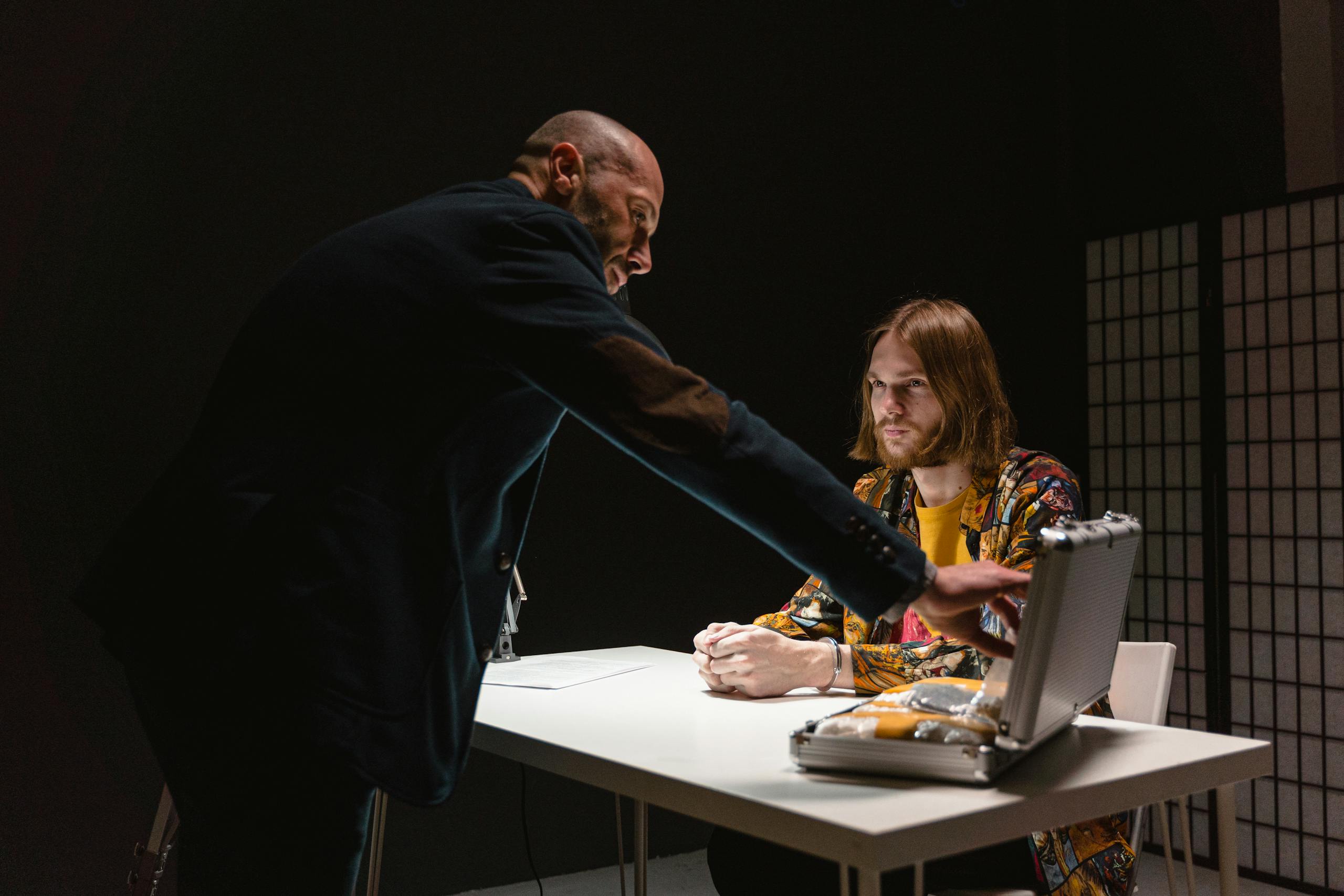 Dramatic interrogation scene with two men discussing a briefcase inside a dark room.