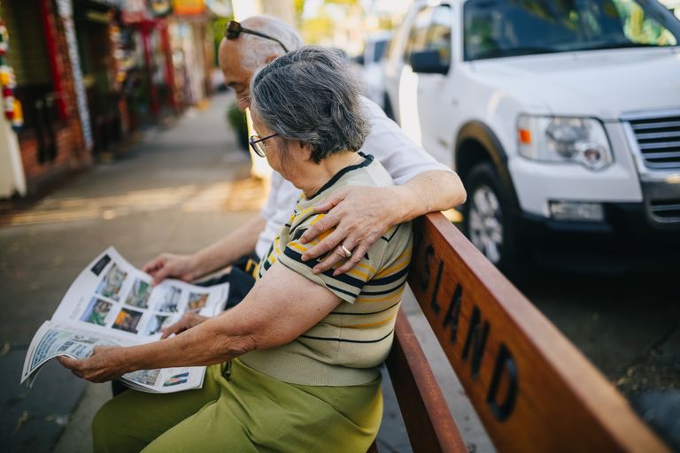 Elderly Asian couple enjoying leisure time on a bench while reading a newspaper outdoors.