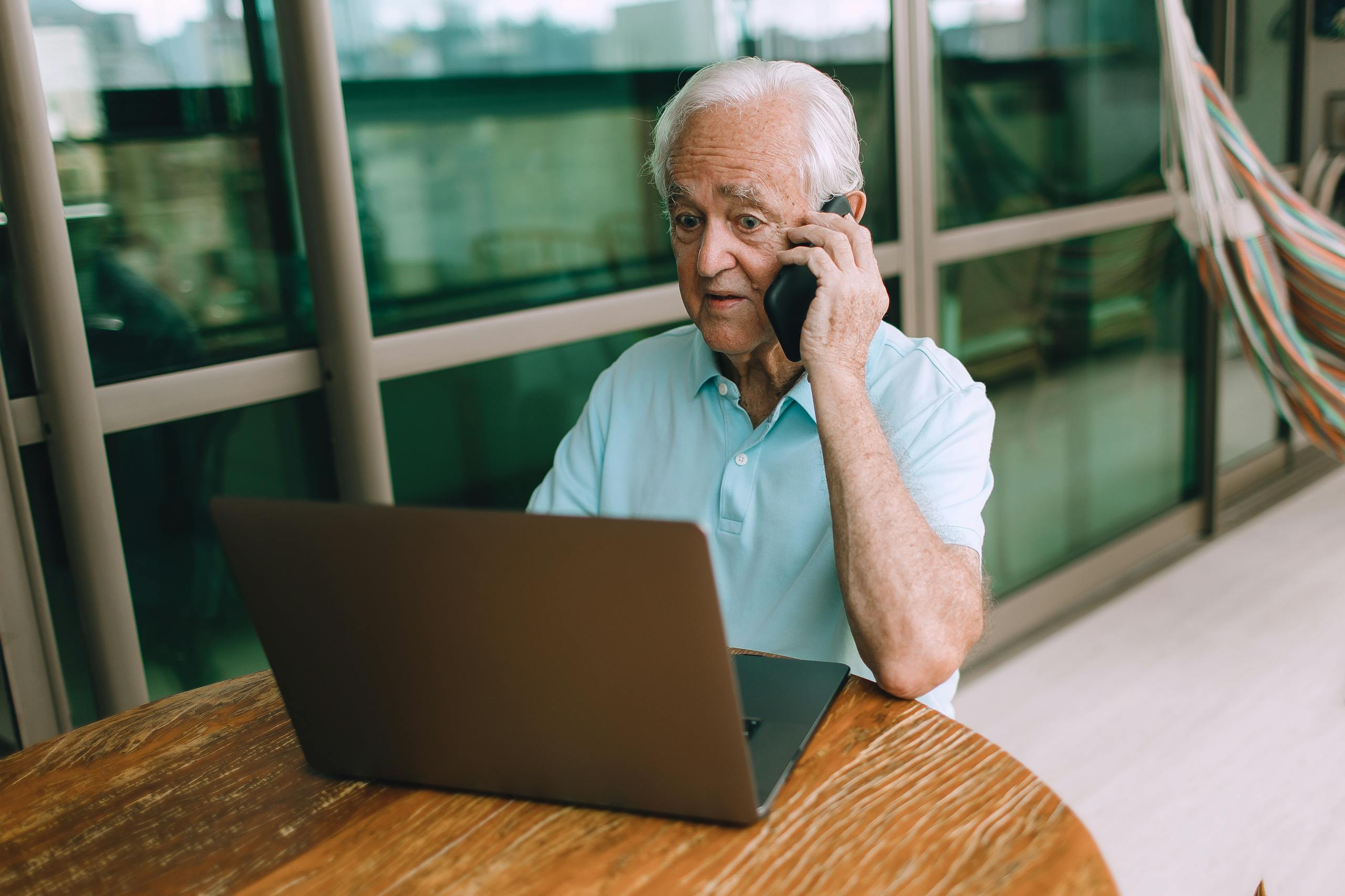 Elderly man with gray hair sitting at table using a laptop and talking on the phone indoors.