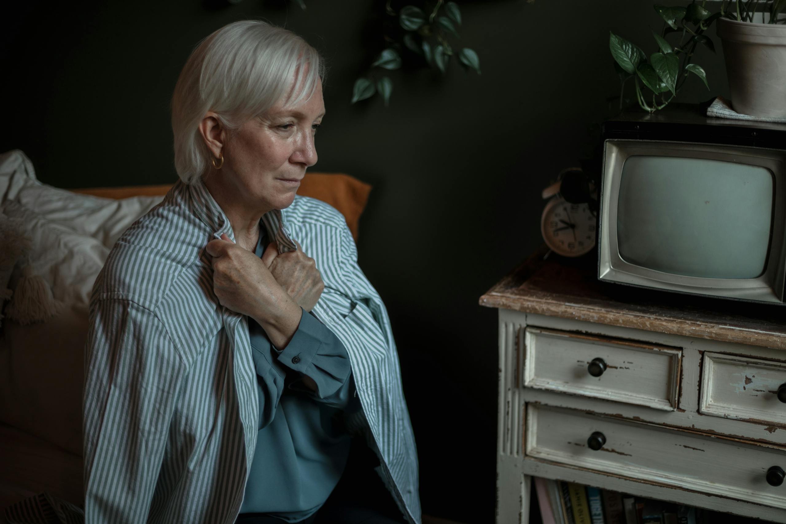 Elderly woman in a striped shirt sitting thoughtfully indoors, clutching her chest.