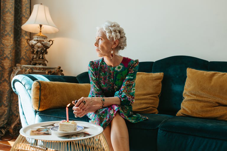 Elegant elderly woman sitting on a couch enjoying cake with a candle indoors. Warm, thoughtful scene.