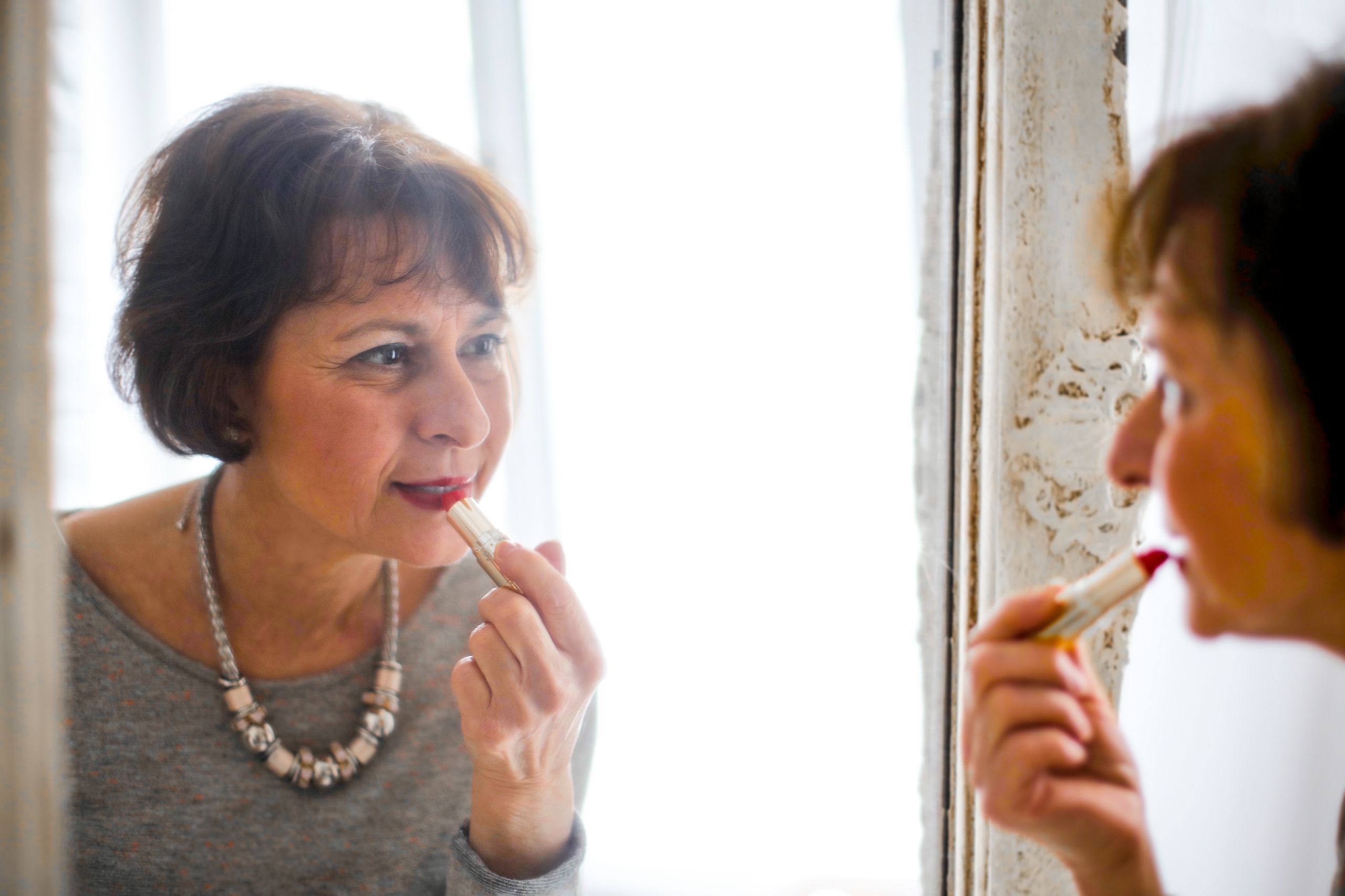 Elegant senior woman applying lipstick while looking in the mirror indoors.