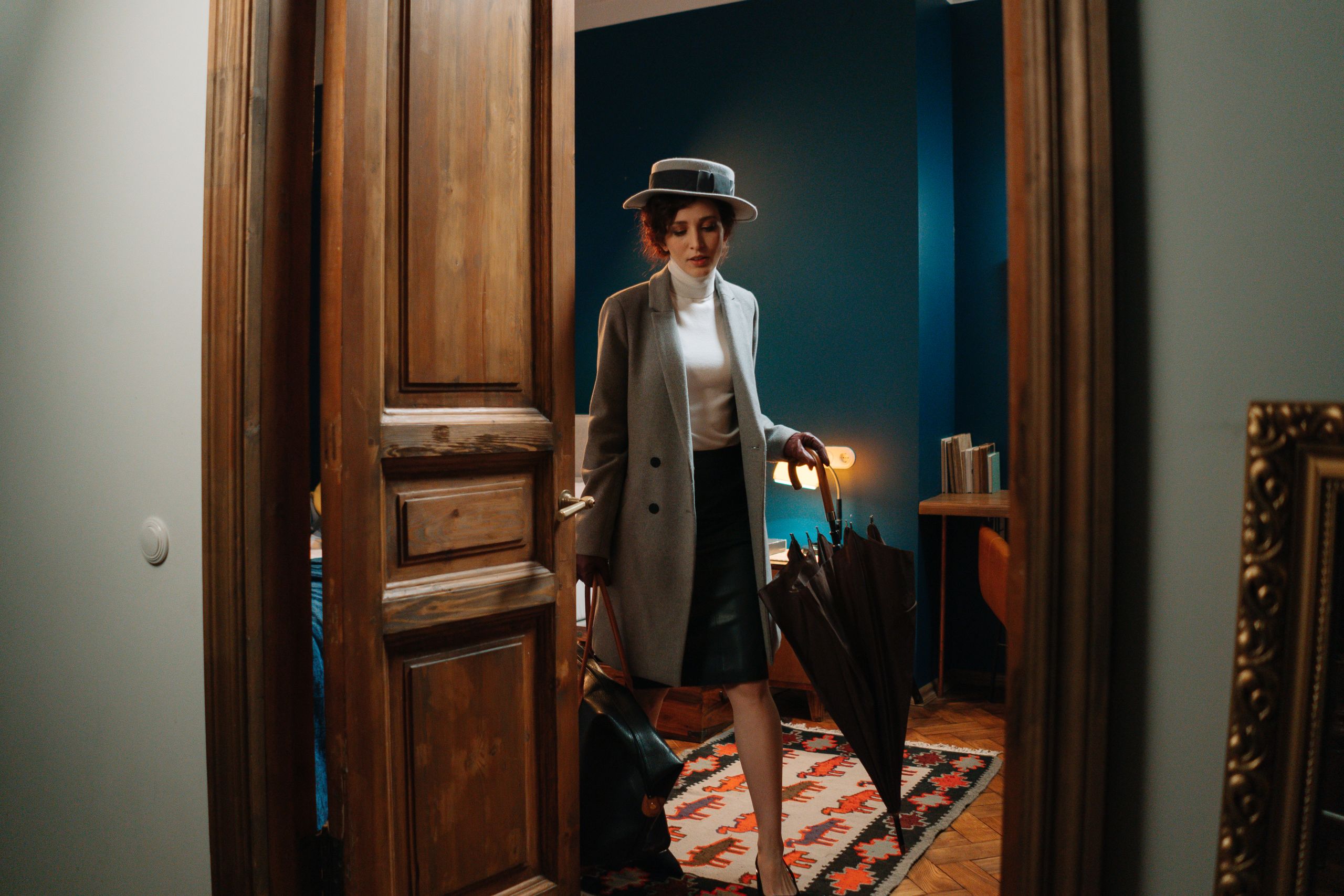 Elegant woman in a hat and coat enters a chic, vintage-inspired room with an umbrella.
