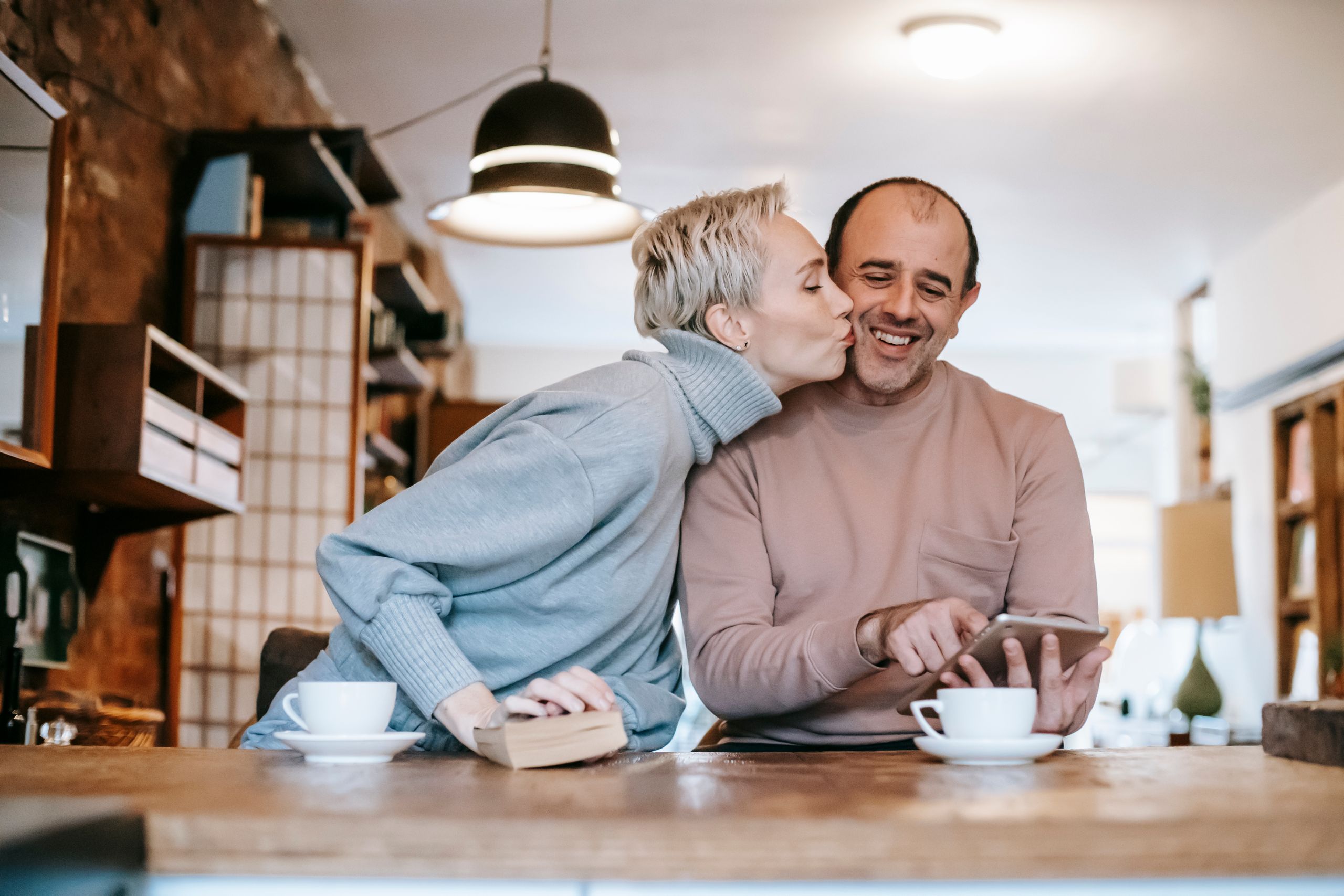 Enamored woman with short blond hair in warm sweater kissing cheek of happy ethnic husband using tablet during breakfast at home