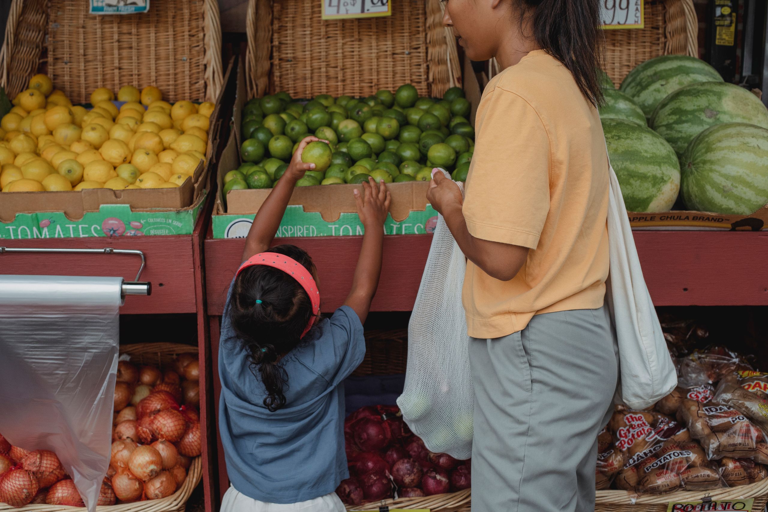 Ethnic girl collecting green apples from box while visiting local market with mother carrying reusable bag