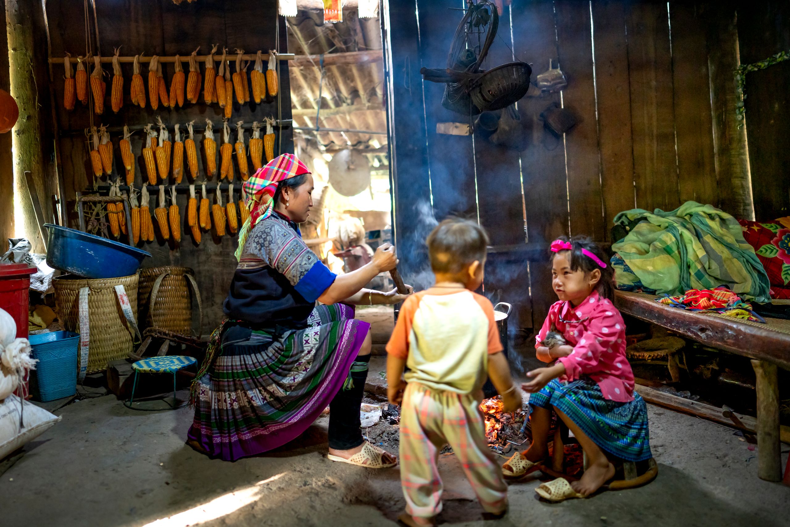 Ethnic mom stirring food in pot against girl speaking with anonymous toddler brother in poor house