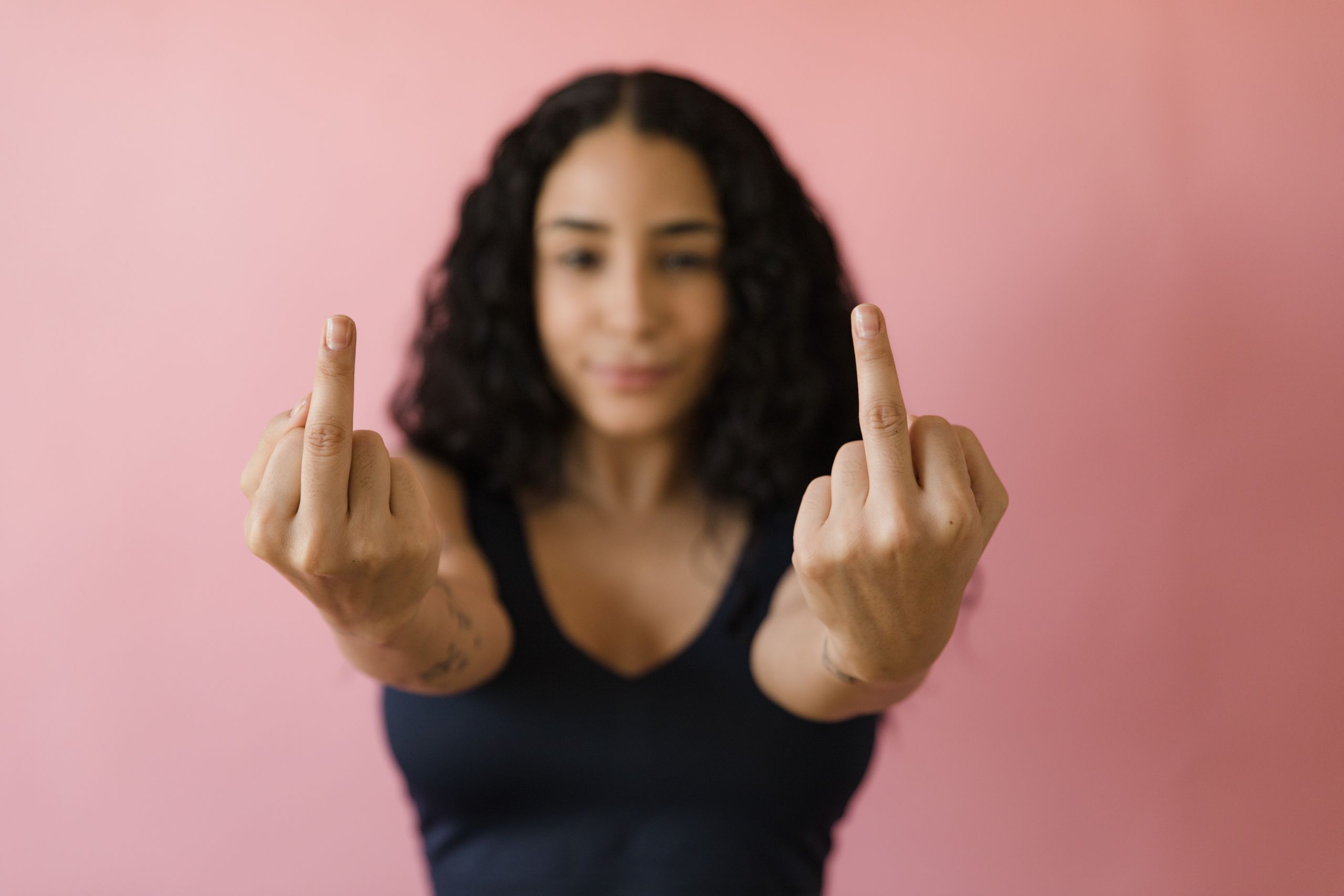 Focus on hands showing middle fingers against blurred pink backdrop, symbolizing defiance.