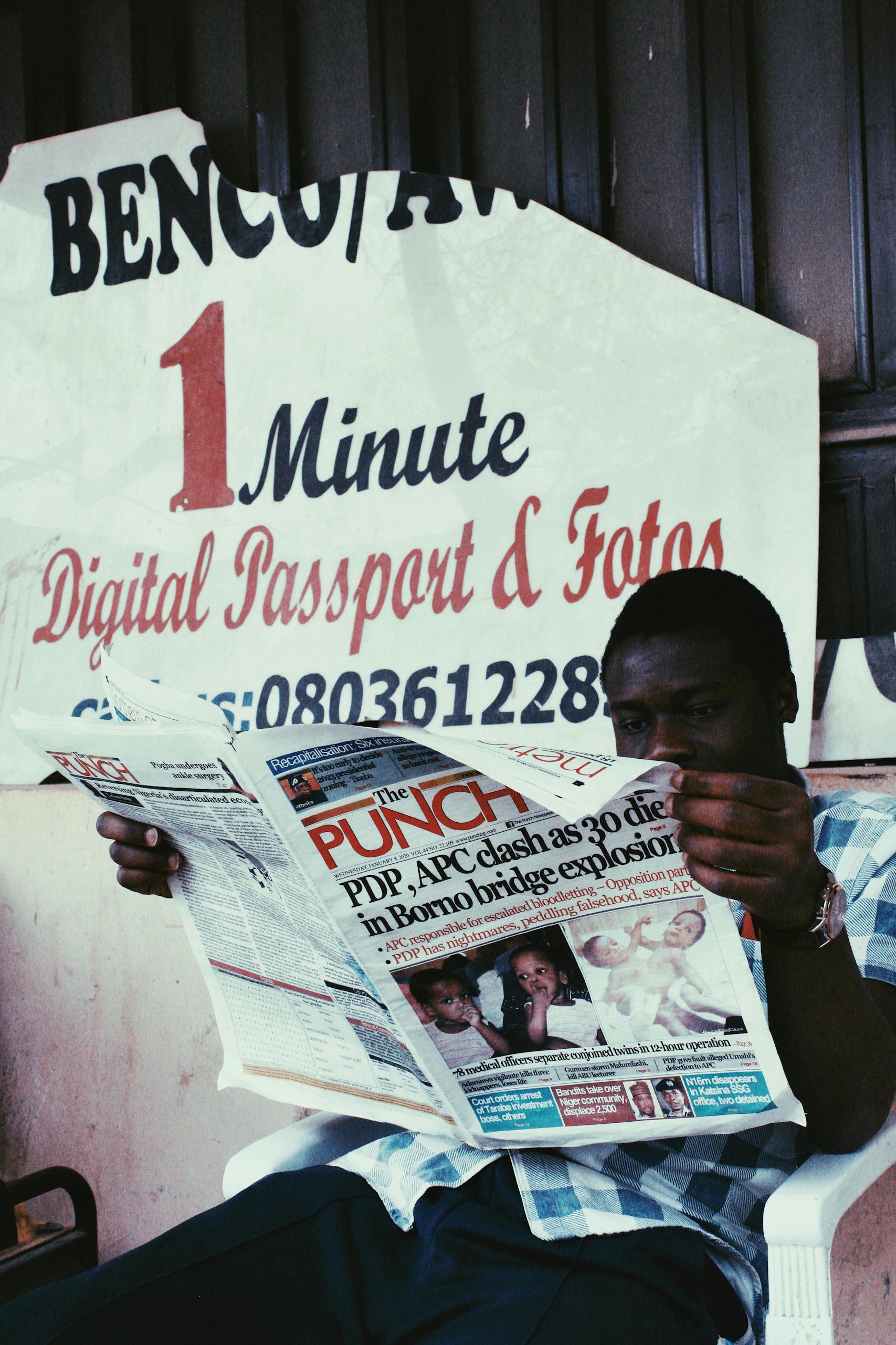 Focused anonymous African American male with newspaper covering half face sitting in chair near wall with torn poster in daylight