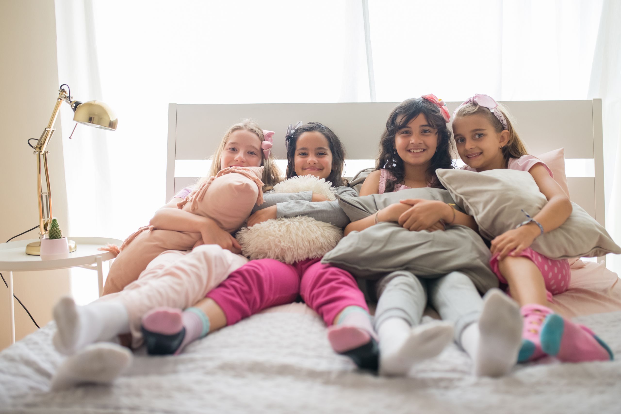 Four cheerful girls enjoying a cozy sleepover party on a bed with pillows and pajamas.