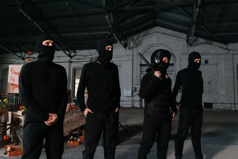 Four individuals in black attire and balaclavas standing in an industrial setting.