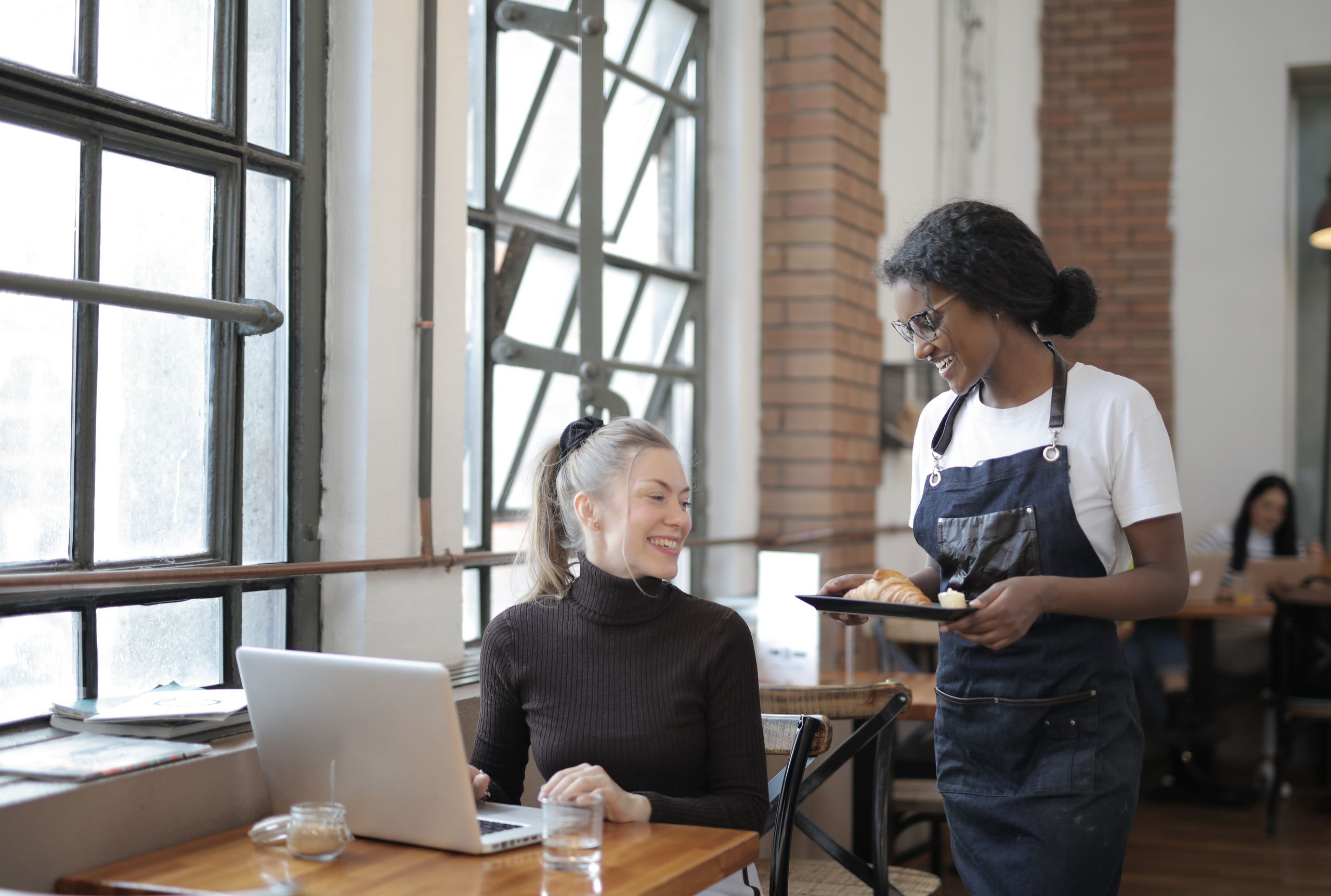 Friendly interaction in a caf&eacute; with a waitress serving a customer using a laptop. Bright and welcoming atmosphere.