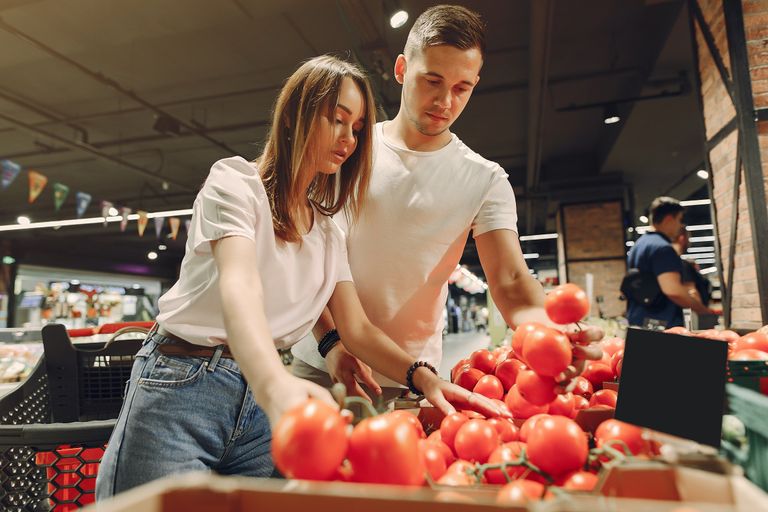 From below of young man and woman in casual clothes standing near counter with vegetables and choosing fresh tomatoes while buying products together in supermarket
