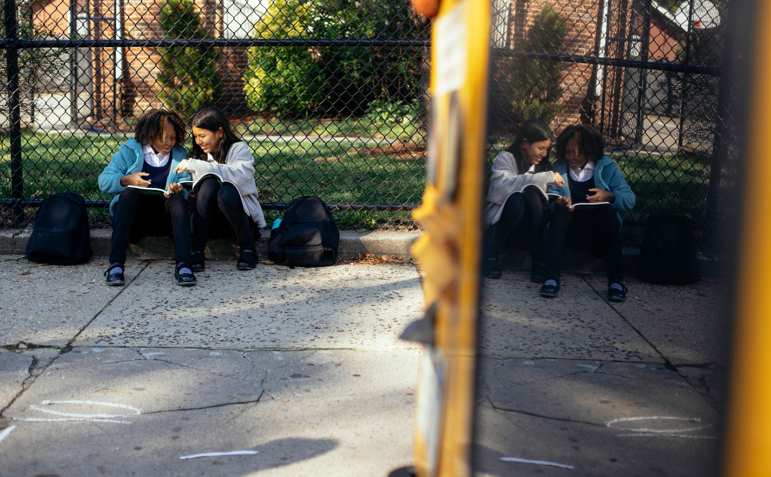 Full body happy multiracial girls classmates with textbooks sitting together on ground near school fence and chatting on sunny autumn weather