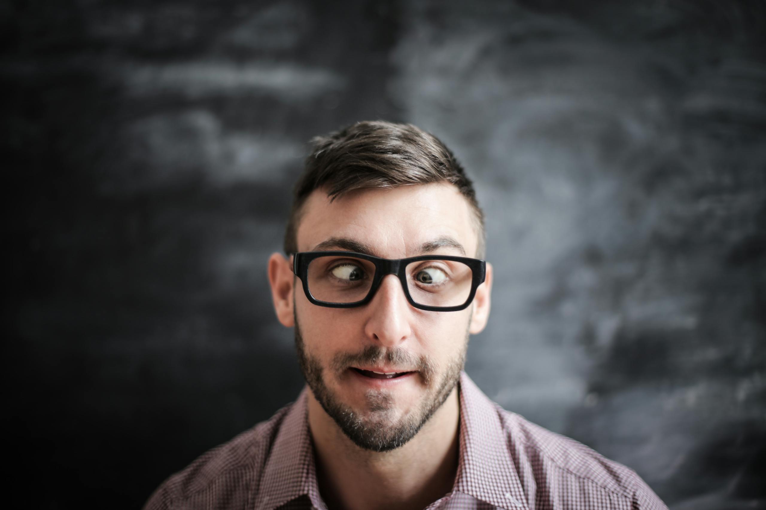 Funny portrait of a man making a goofy expression, wearing glasses against a chalkboard background.