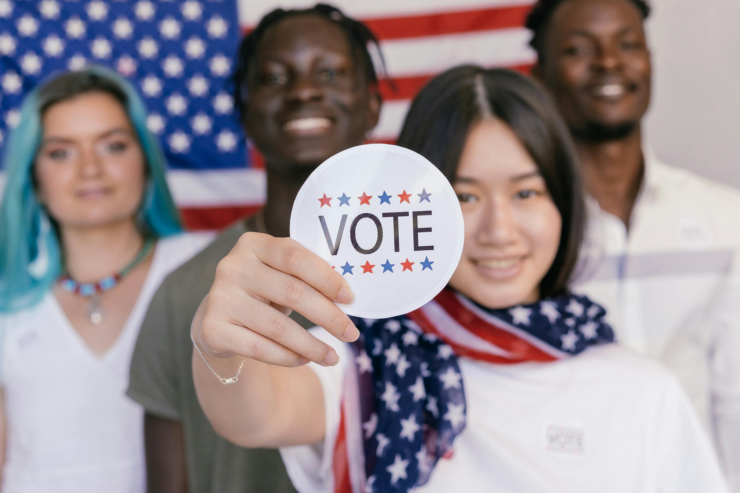 Group of diverse young adults promoting voting with a focus on a 'VOTE' sticker.