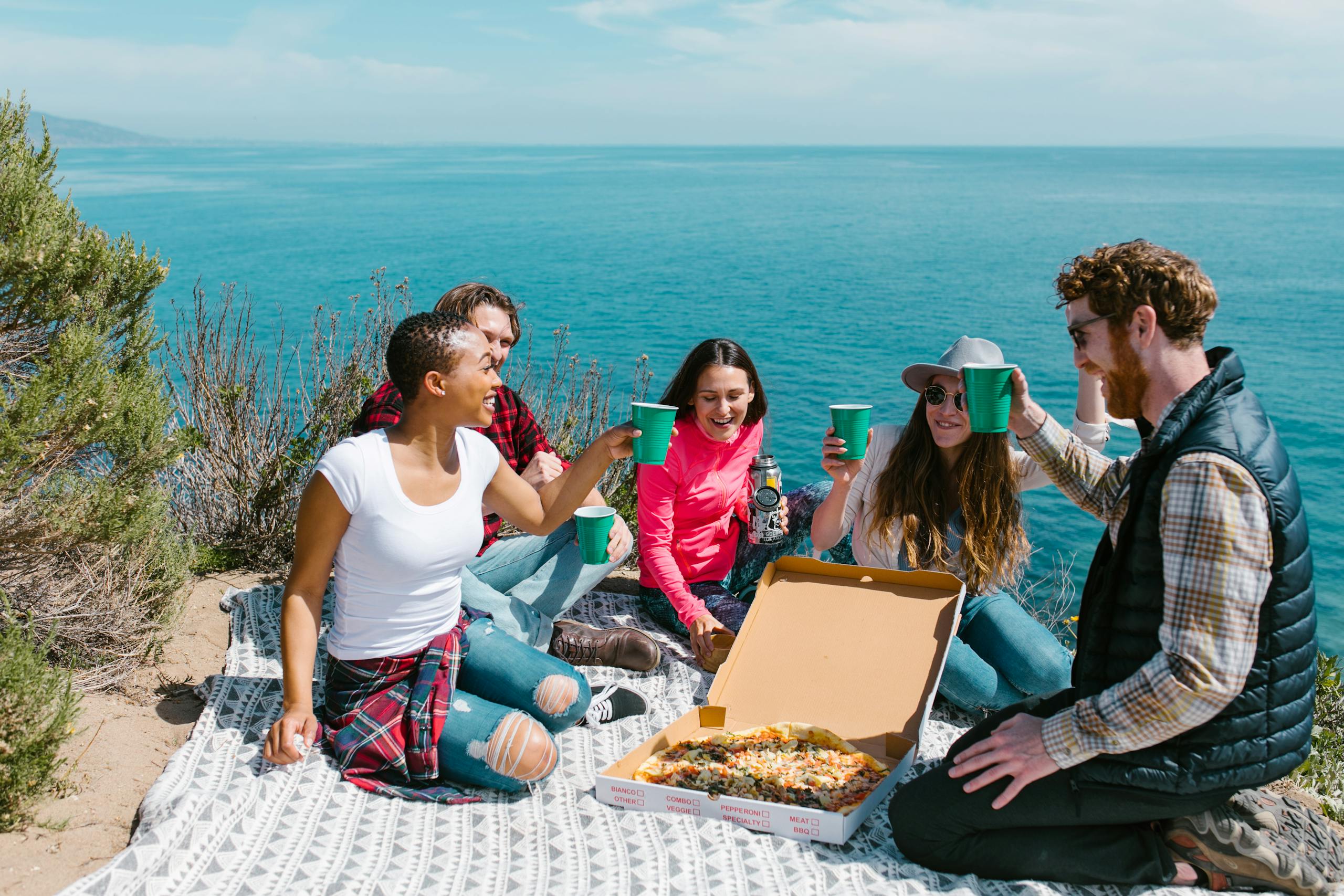 Group of friends enjoying a picnic by the seaside with pizza and drinks on a sunny day.