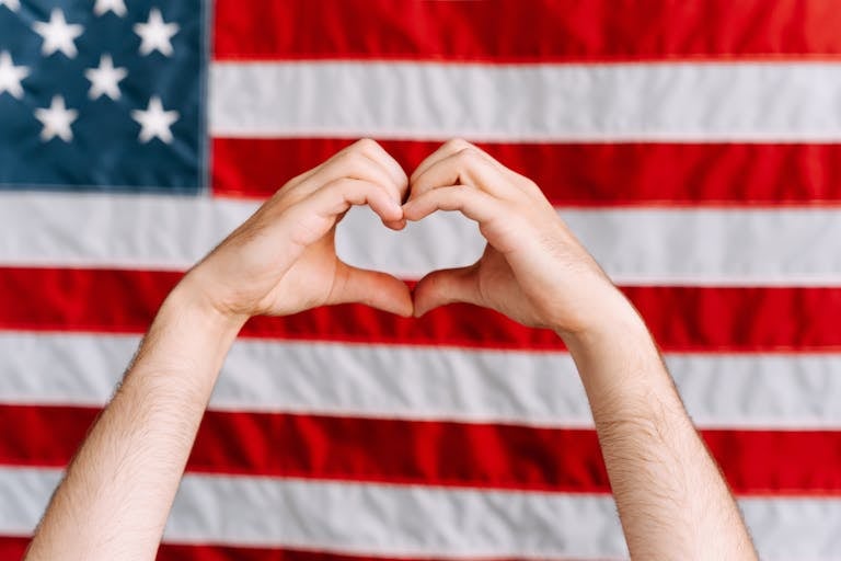 Hands forming a heart shape in front of the American flag, symbolizing love and patriotism.