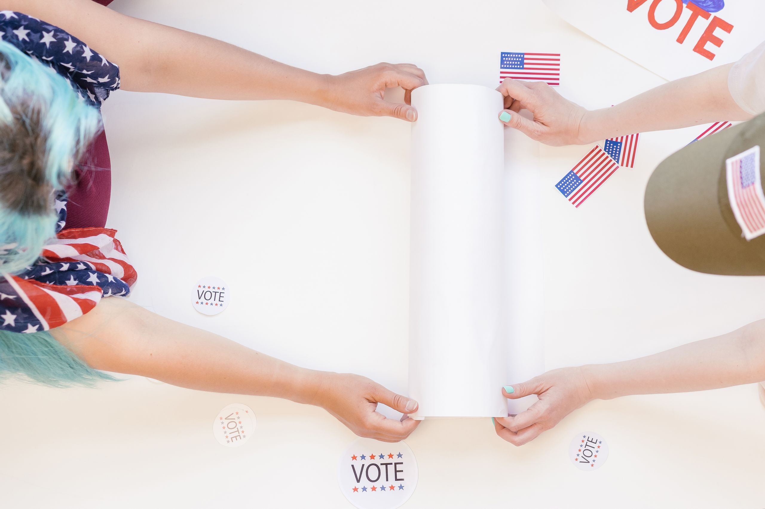 Hands of people holding voting materials, American flags, and 'vote' stickers on a white table.