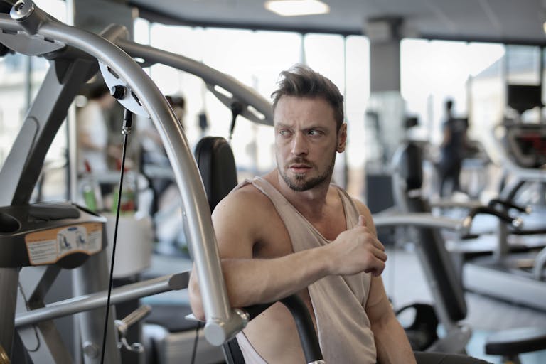 Handsome male athlete in sportswear sitting in modern fitness center and resting after training while looking away