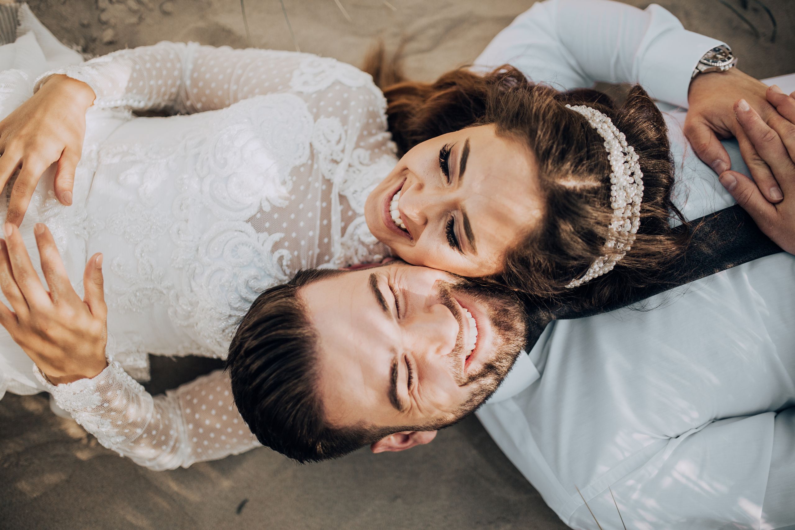 Happy bride and groom lying side by side on the beach in wedding attire. Capturing a joyful and intimate moment.