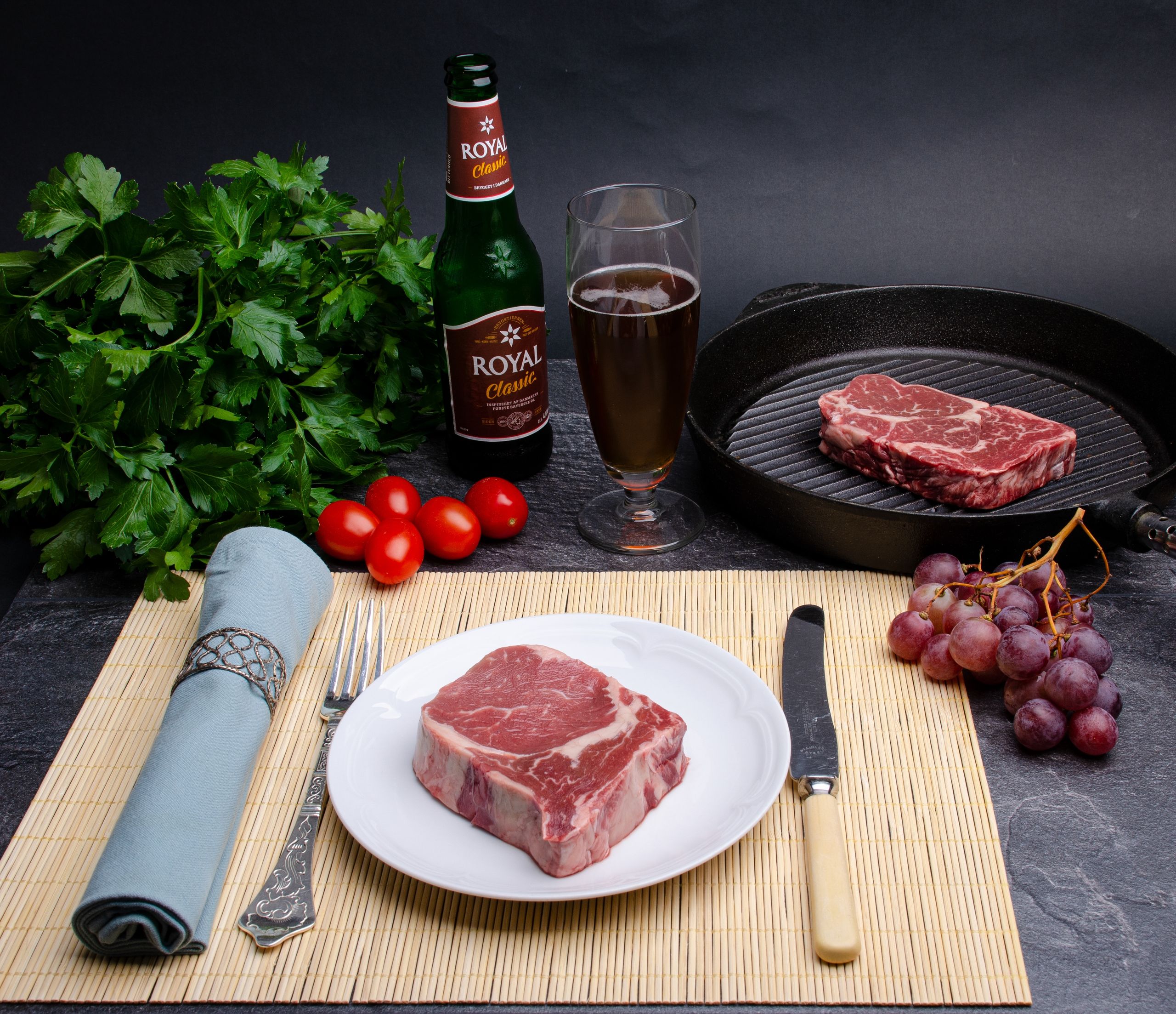 High angle of slices of raw meat on plate and grill pan served with bottle of beer and grapes with herbs