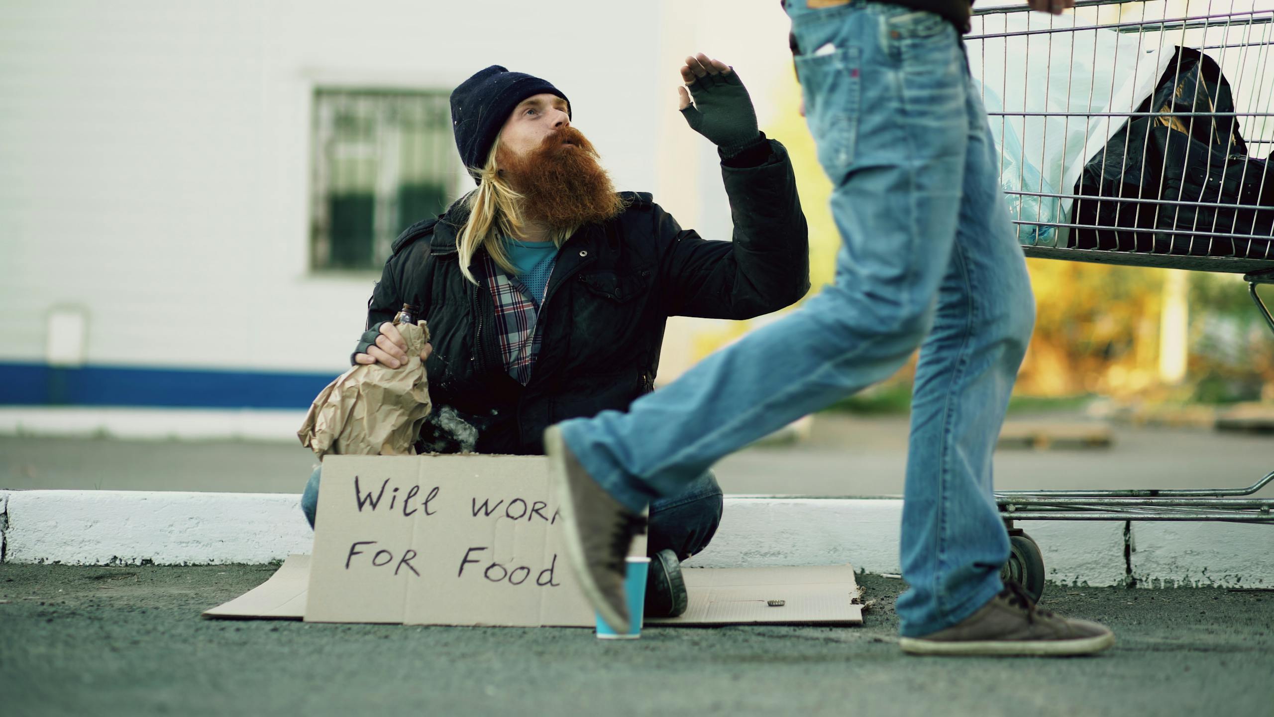 A homeless man is sitting beside a shopping cart holding a sign reading 'Will Work For Food.'