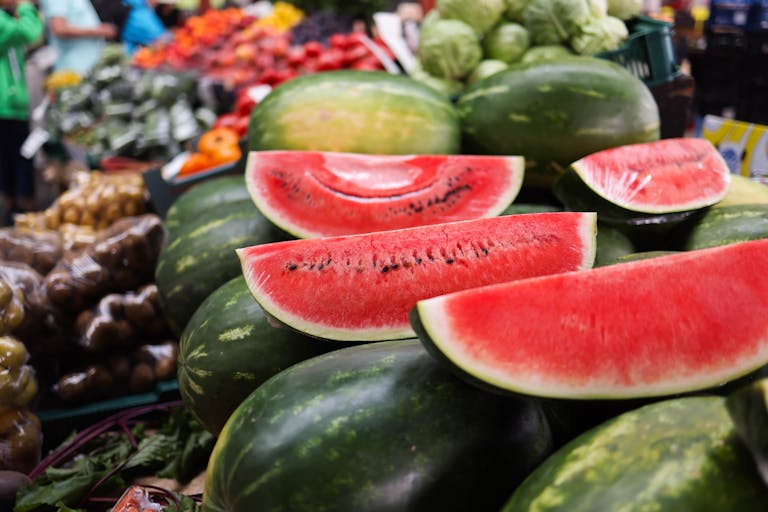 Juicy watermelon slices stacked among fresh fruits and vegetables at an outdoor market.