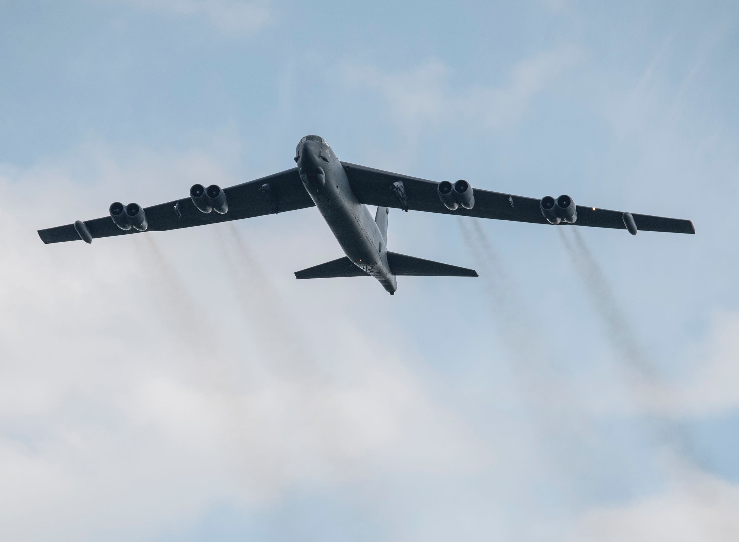 Large military aircraft flying through clear sky, emitting smoke trails from engines.