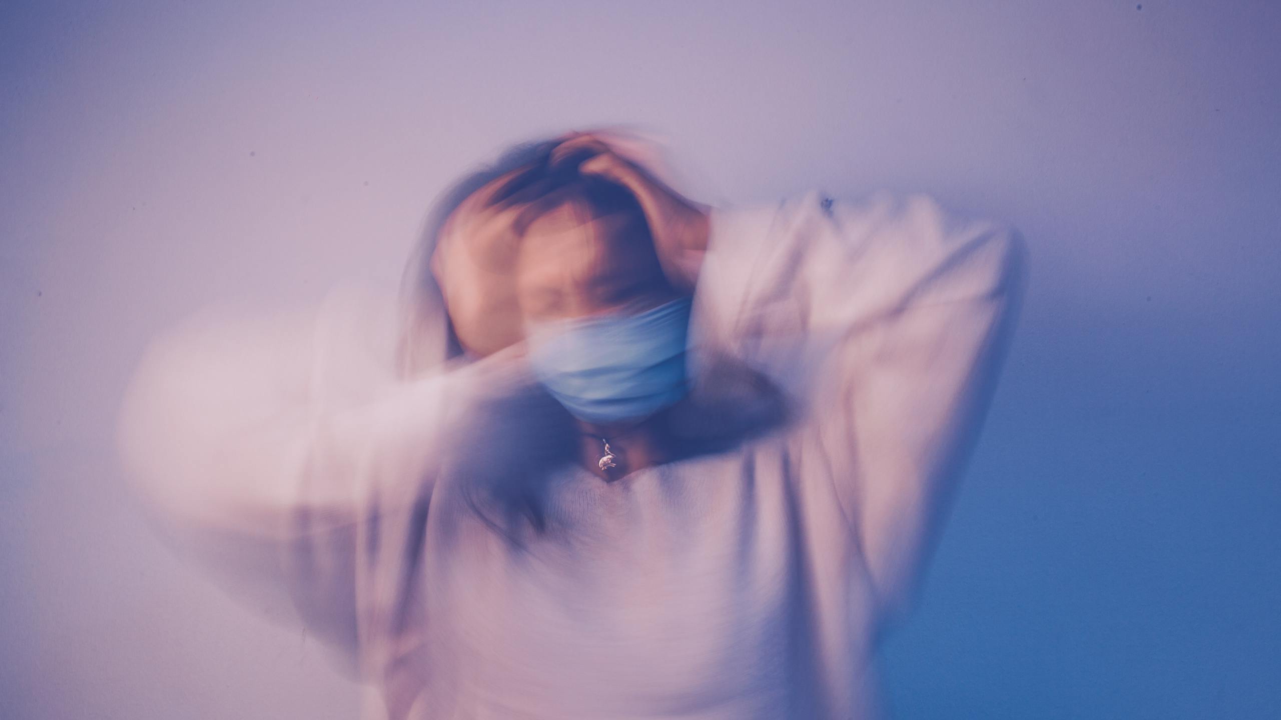Long exposure concerned puzzled female wearing protective face mask standing against blue wall and touching head in frustration