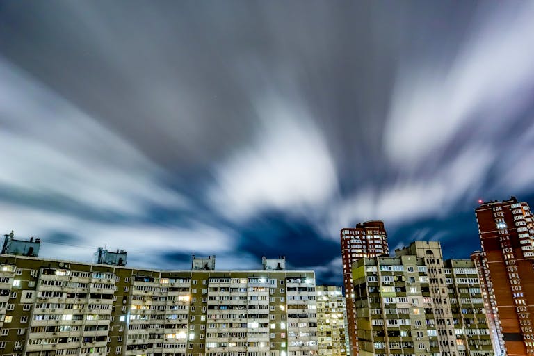 Long exposure of urban skyline with illuminated buildings and fast-moving clouds creating a dynamic city night scene.