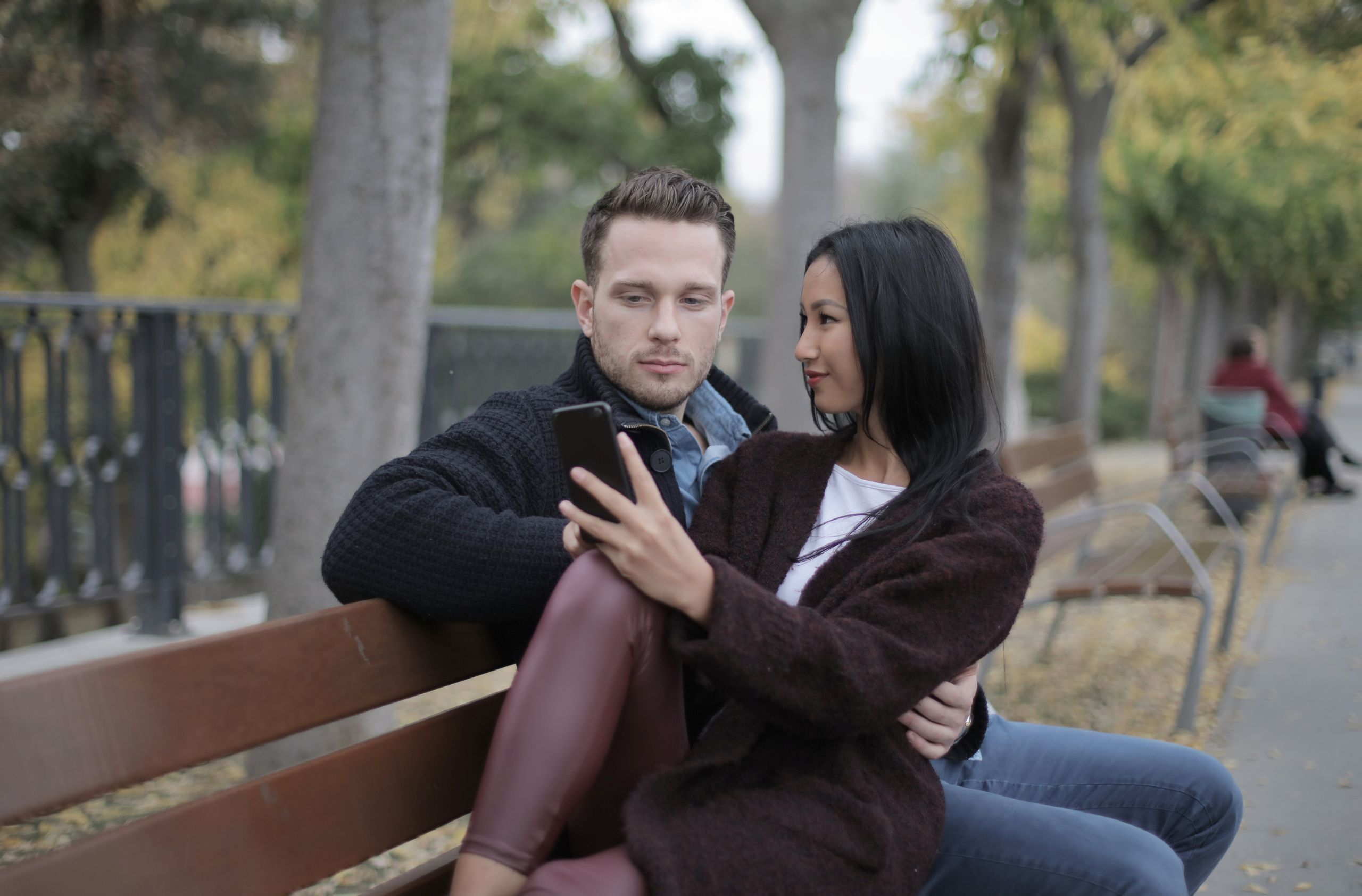 Loving modern multiracial couple in casual warm clothes sitting together on bench and using smartphone while spending time in park in city