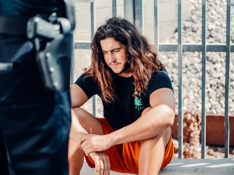 Man in black shirt with wavy hair sitting outdoors by a metal fence, looking contemplative.