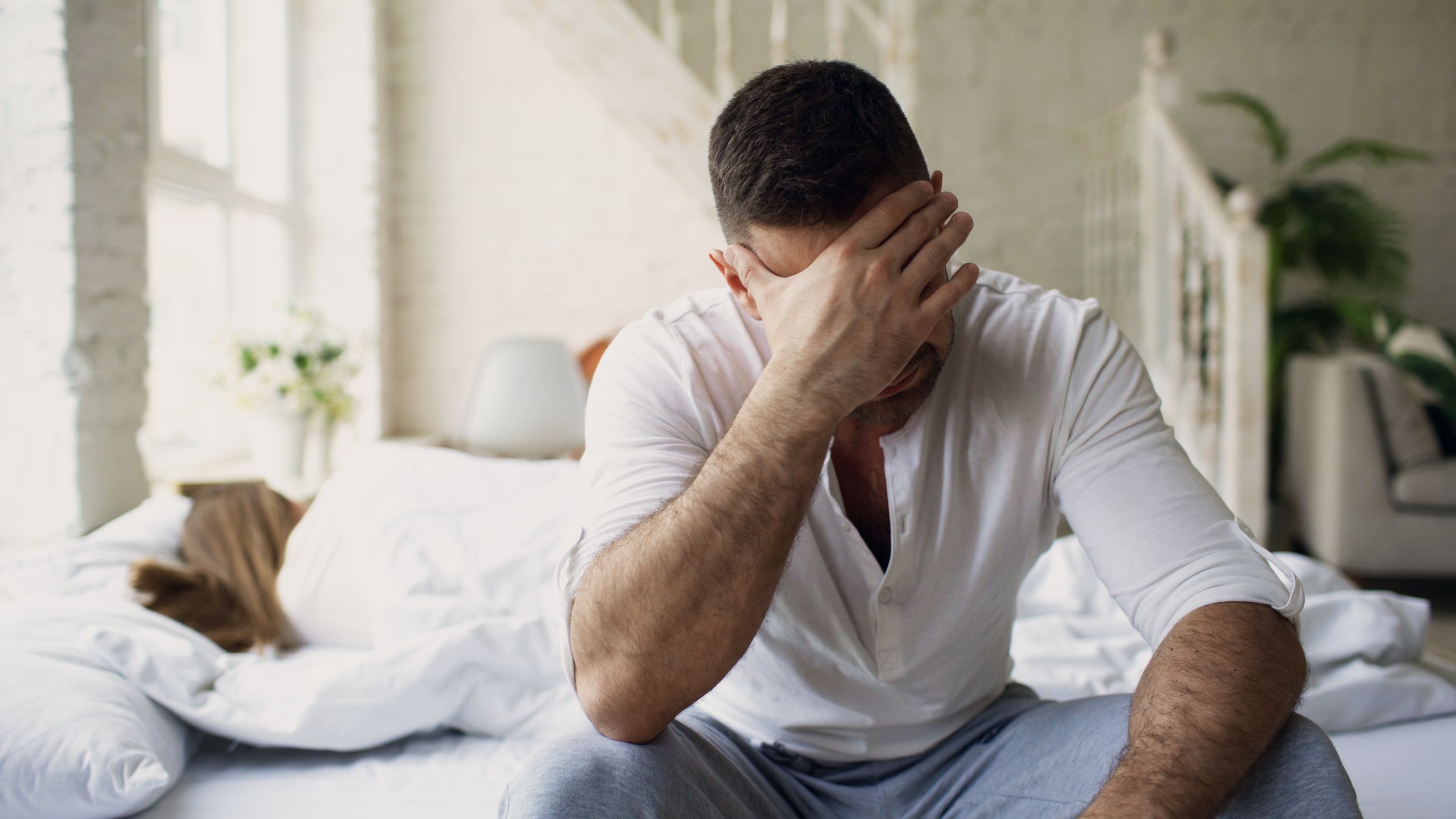 Man in distress sitting on bed in sunlit room, hand on face.
