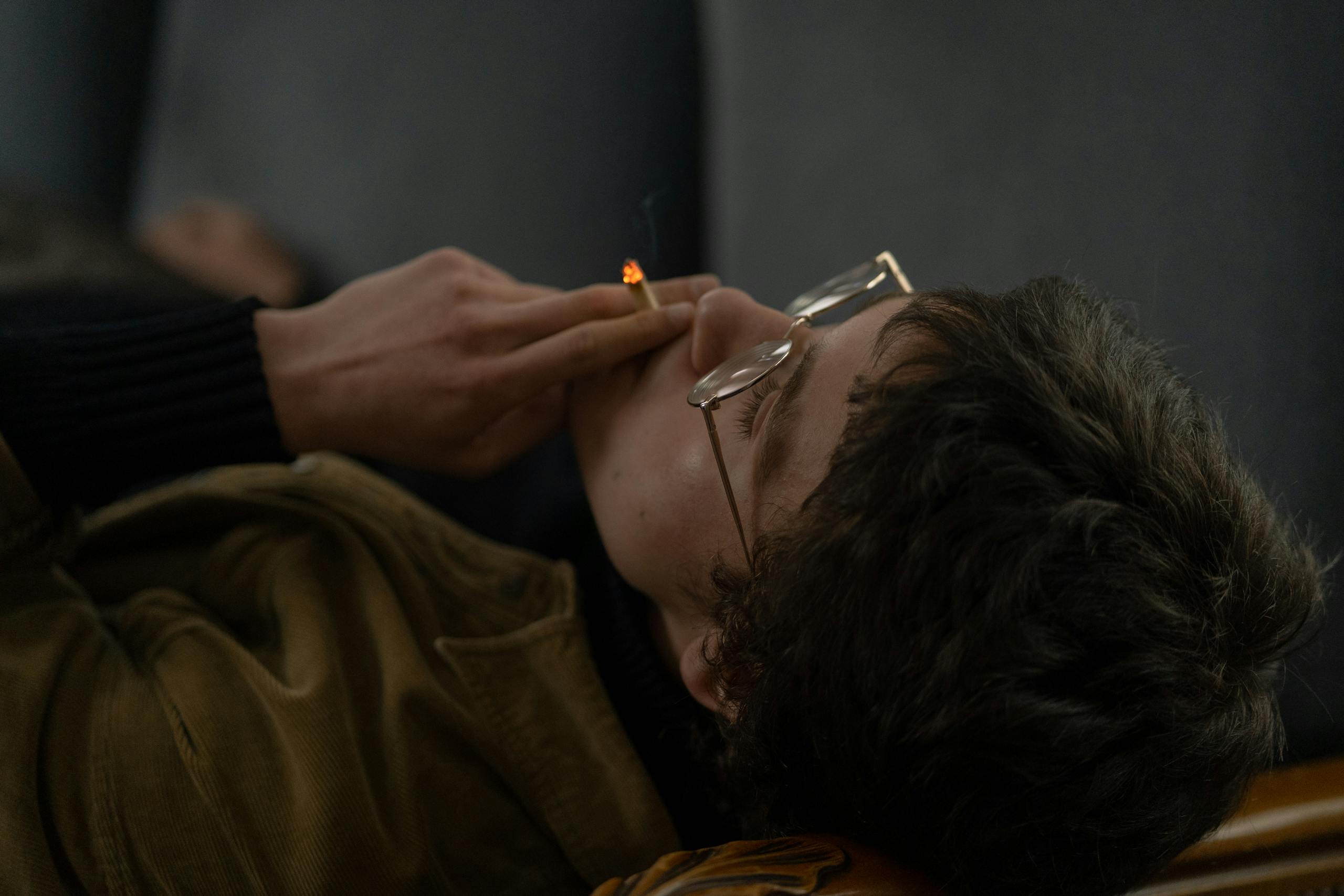 Man in glasses and brown jacket relaxing indoors while smoking. Moody atmosphere.